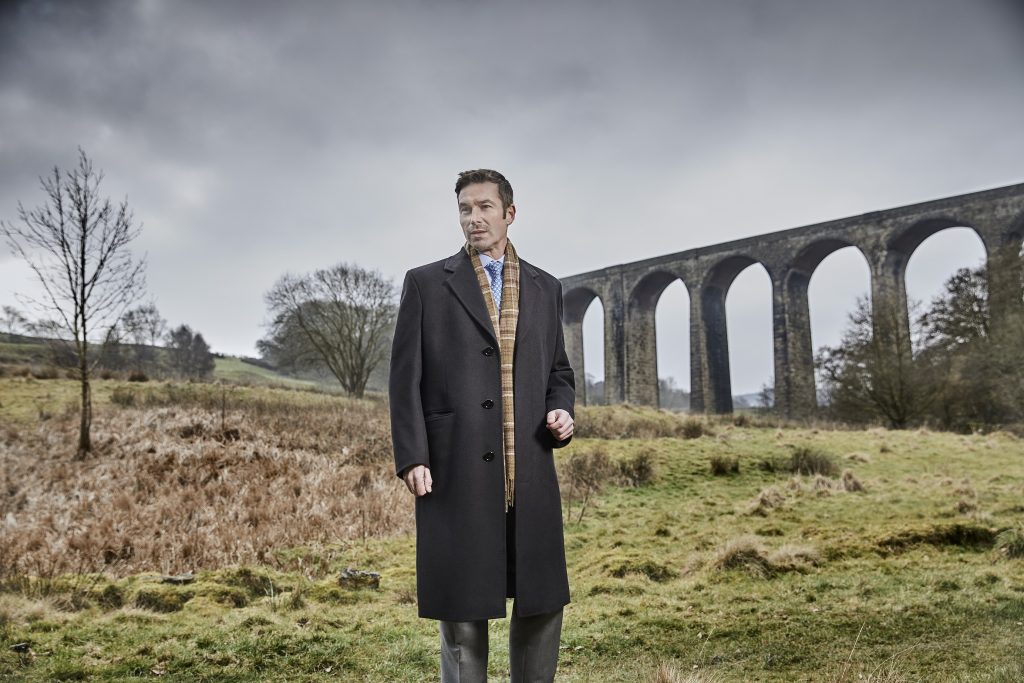 A male model stands in from of a Yorkshire landscape of grassy hills and a viaduct wearing a smart grey coat made from Standeven's Everest Autumn/ Winter cloths